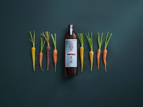 Still life flat lay composition of raw carrots with green tops and a bottle of vegetable juice on a dark teal background.
