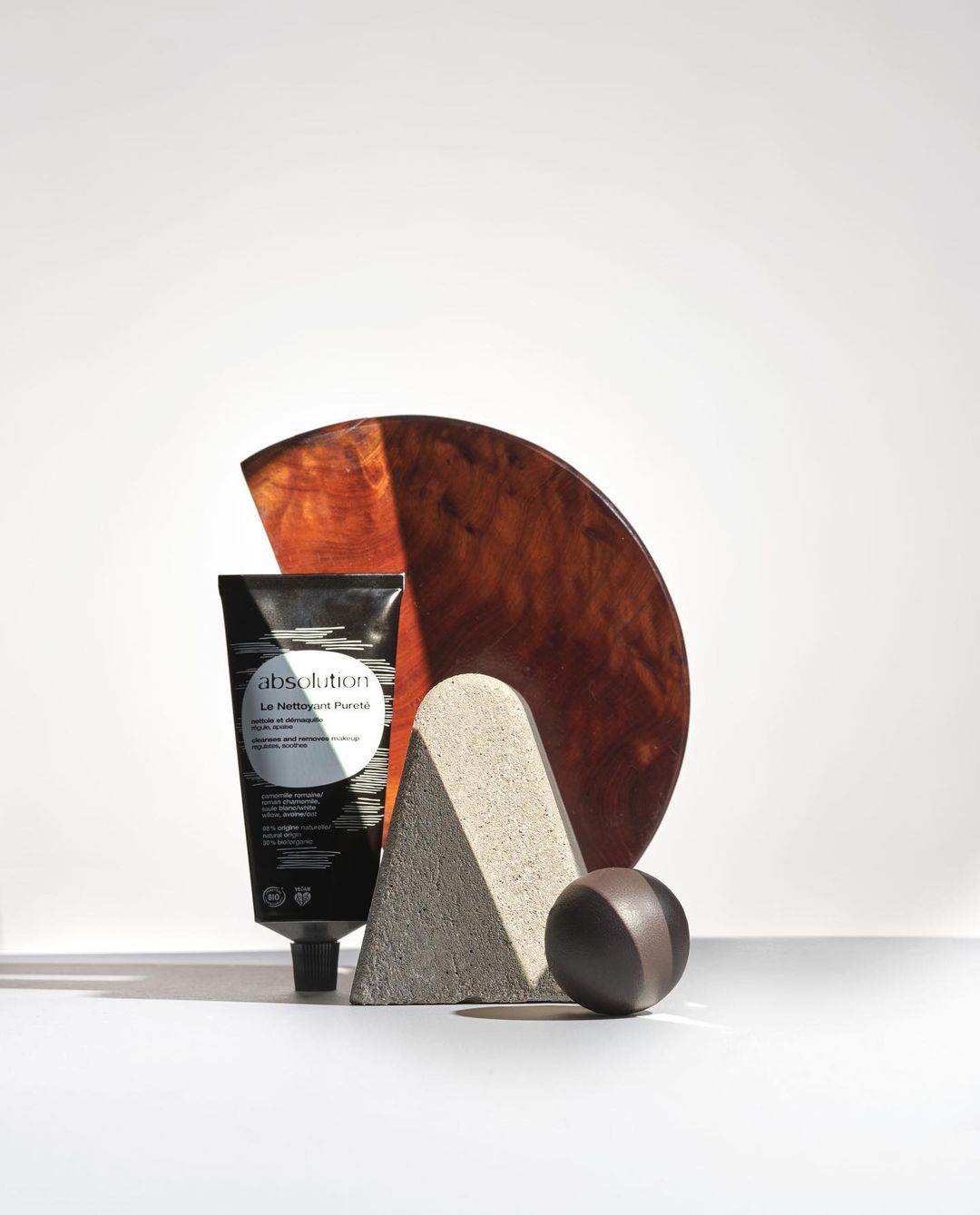 Black tube of Absolution Le Meilleur Masque rests beside brown wooden semi-circle, gray geometric stone, and brown sphere against white.
