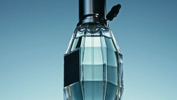 Close-up studio shot of a faceted glass perfume bottle with a black cap against a light blue background. The bottle is the primary subject.