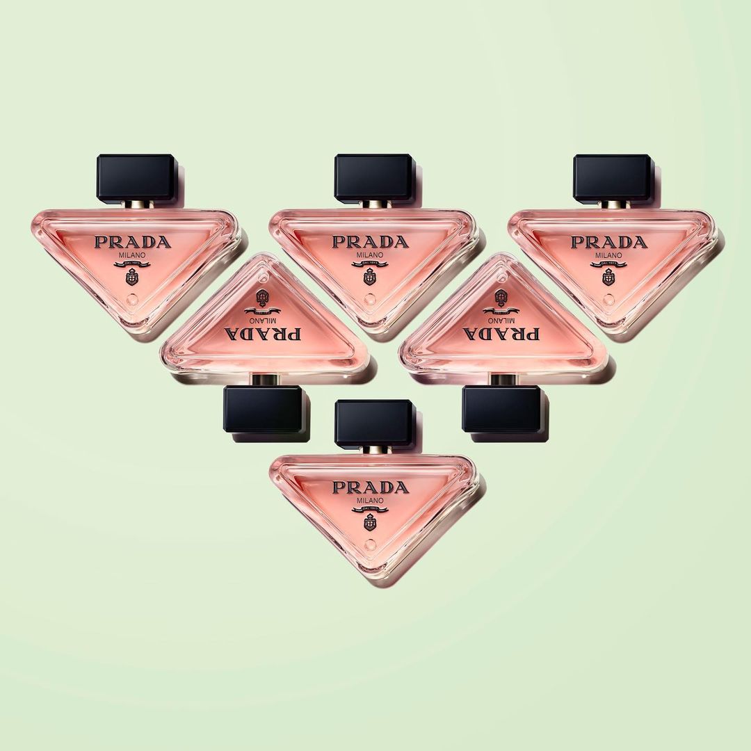 Arrangement of six Prada Paradoxe perfume bottles on a soft, light green background, showcasing their triangular shape and branding.