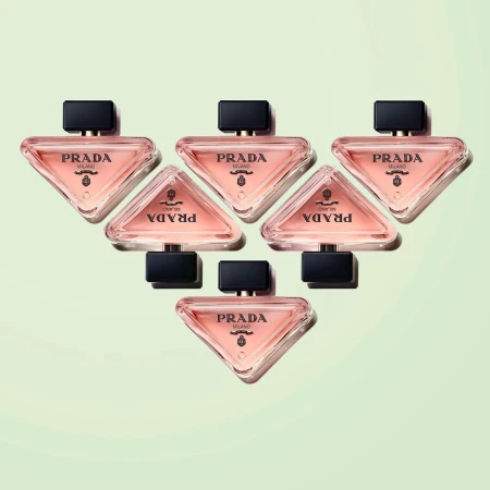 Arrangement of six Prada Paradoxe perfume bottles on a soft, light green background, showcasing their triangular shape and branding.