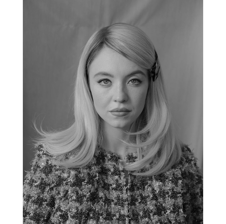 Black and white portrait of Sydney Sweeney with blonde hair, a textured jacket, and a hair clip against a blurred grey background.