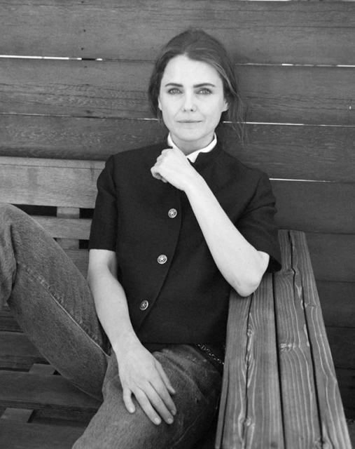 Black and white portrait of an adult woman in jeans and a jacket, seated on a wooden bench. Her hand is to her chin.