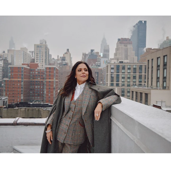 Adult woman wearing a patterned suit and overcoat posing on a rooftop with a cityscape in the background on an overcast day.