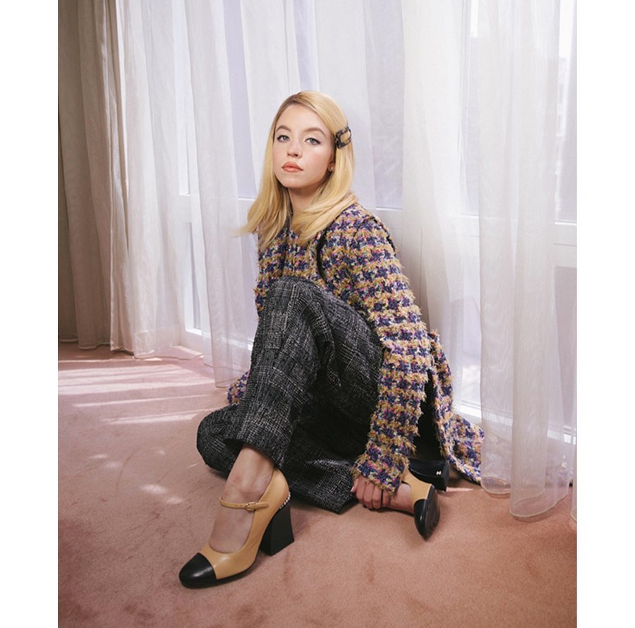 A woman with blonde hair sits on the floor in a tweed jacket and wide-leg pants, wearing Mary Jane shoes in a brightly lit room with sheer curtains.