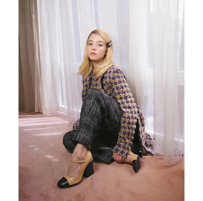 A woman with blonde hair sits on the floor in a tweed jacket and wide-leg pants, wearing Mary Jane shoes in a brightly lit room with sheer curtains.