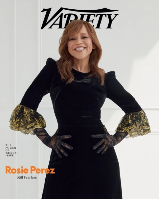 Rosie Perez in black velvet dress with lace details on magazine cover, smiling with hands on hips against a white wall.
