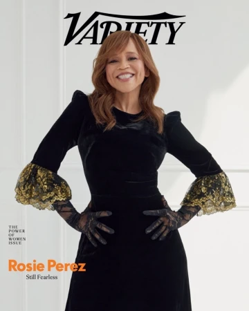 Rosie Perez in black velvet dress with lace details on magazine cover, smiling with hands on hips against a white wall.