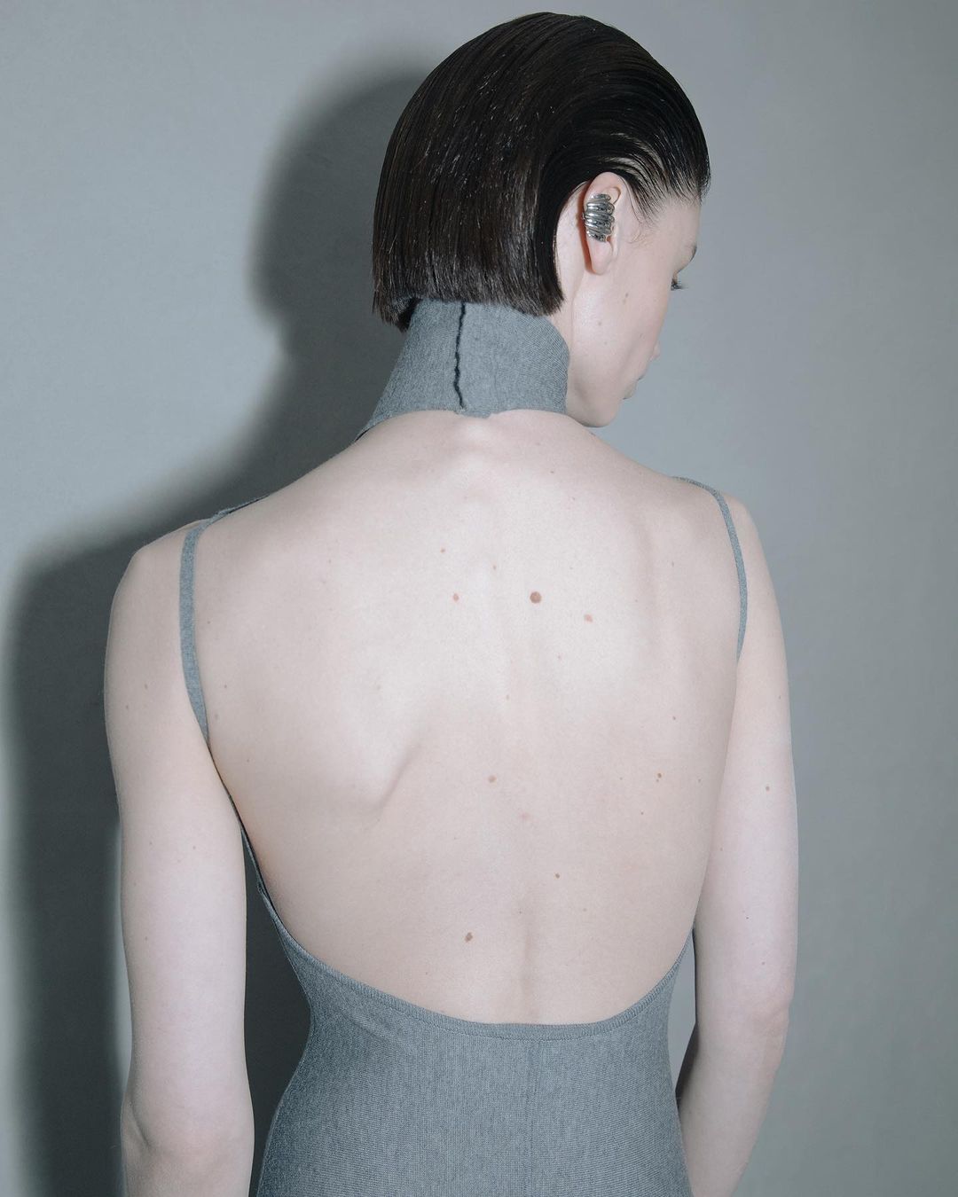 Back view of a woman with short, slicked-back hair wearing a gray, open-back dress and an ear cuff against a gray backdrop.