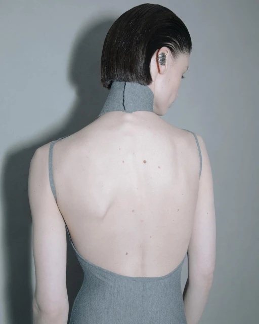 Back view of a woman with short, slicked-back hair wearing a gray, open-back dress and an ear cuff against a gray backdrop.