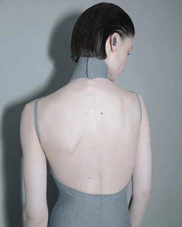 Back view of a woman with short, slicked-back hair wearing a gray, open-back dress and an ear cuff against a gray backdrop.
