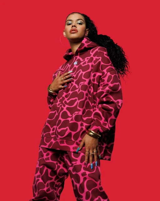 Portrait of a young woman wearing a patterned hoodie and pants against a red backdrop. She has gold jewelry and long dark hair.