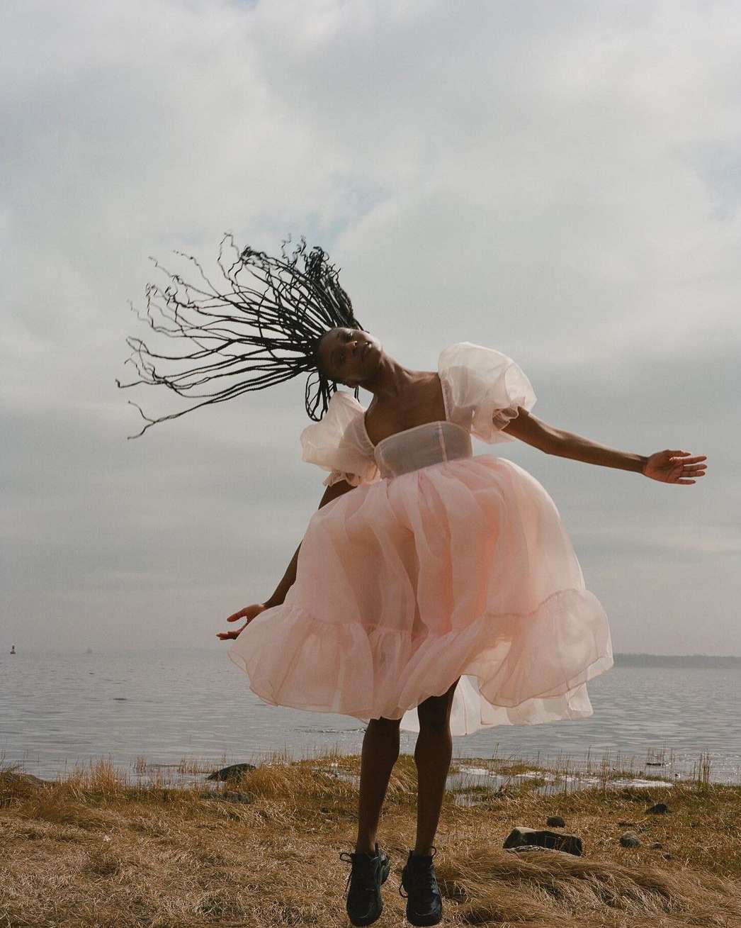 A young adult female model in a pink tulle dress leaps in the air on coastal grasses against an overcast sky by the sea.