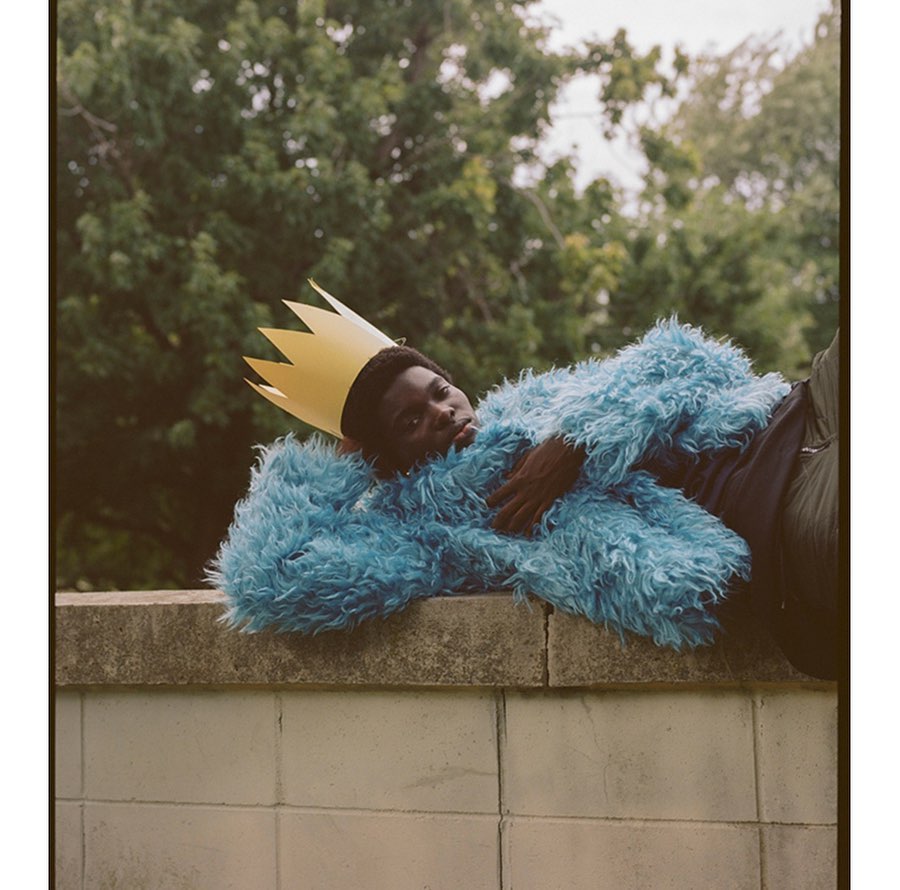 Portrait of a Black adult in a turquoise fur coat and paper crown reclining against a concrete wall surrounded by green trees.