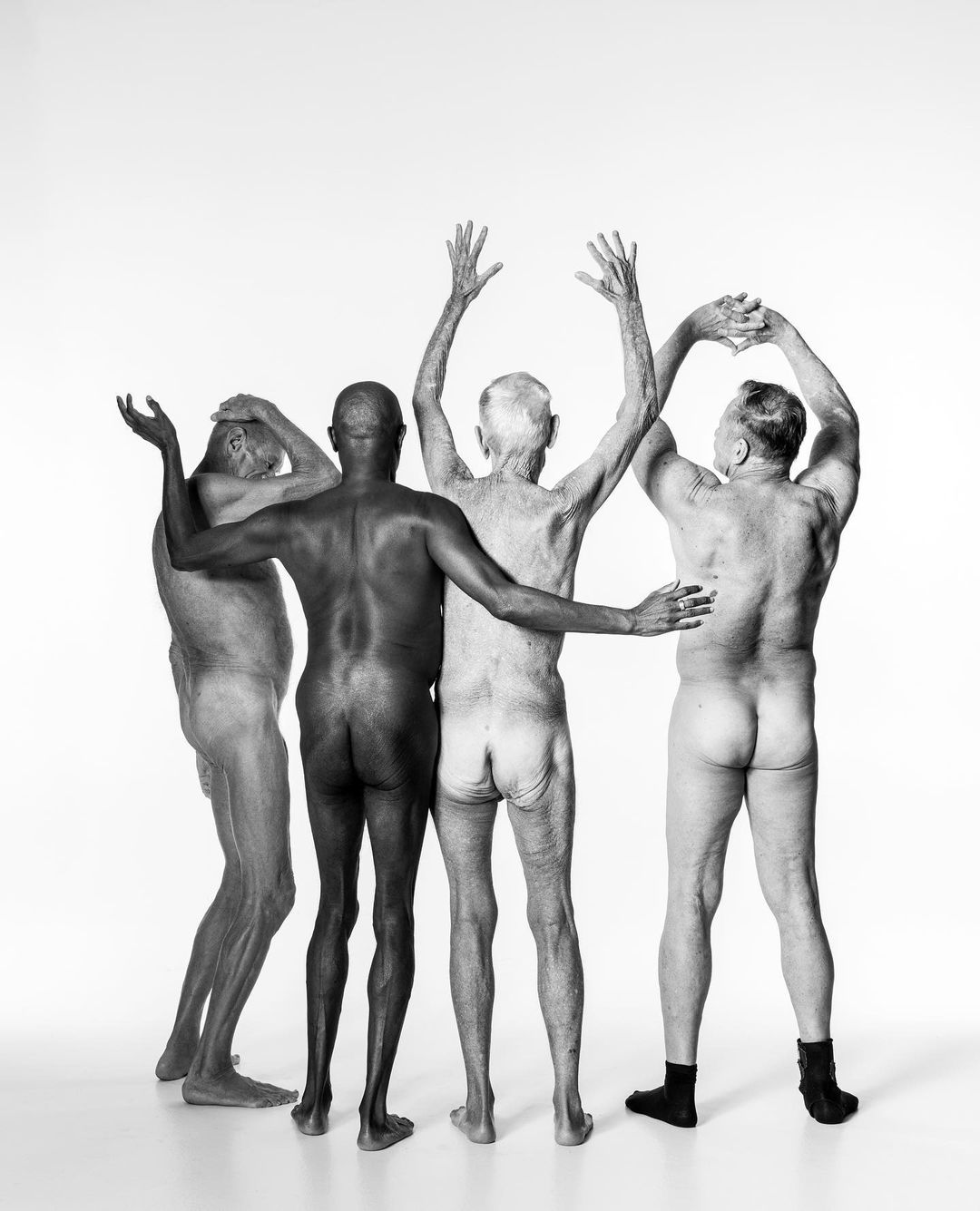 Black-and-white image of four older men standing in a line with their arms raised against a bright white background in a photo studio.