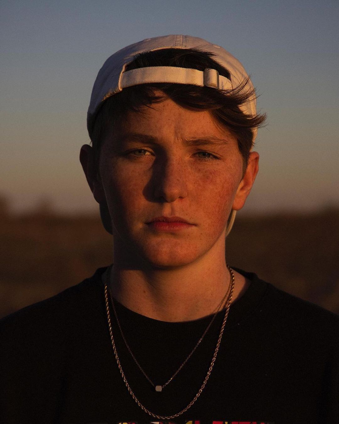 Portrait of a young adult with a cap and layered necklaces, posing outdoors at sunset, bathed in warm, golden light against a blurry background.