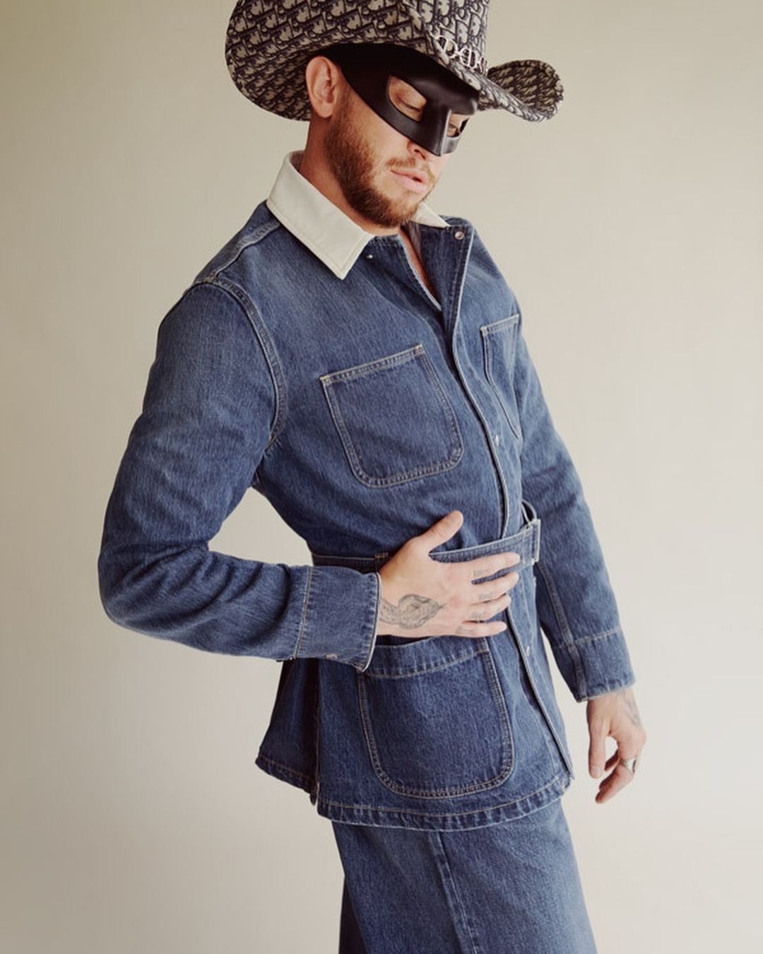 Man in denim jacket and skirt wearing a patterned cowboy hat and mask, standing in front of a light beige background in a studio setting.