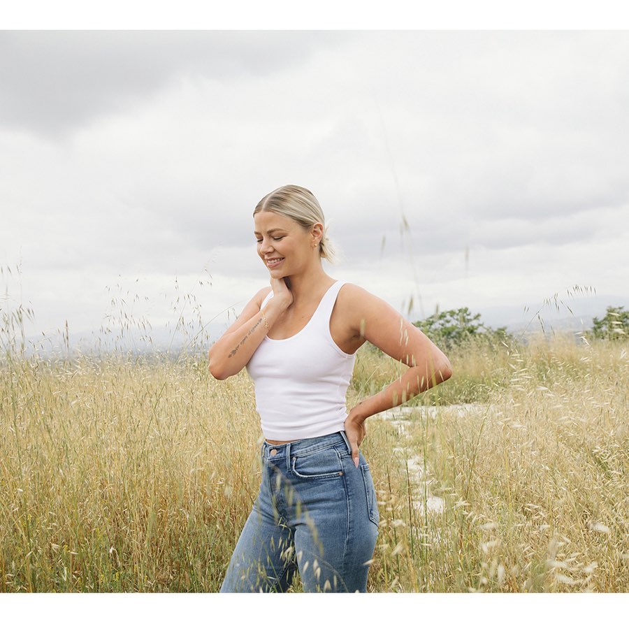 Young woman in a white tank top and blue jeans smiles while standing in a golden field beneath an overcast sky, portrait shot.