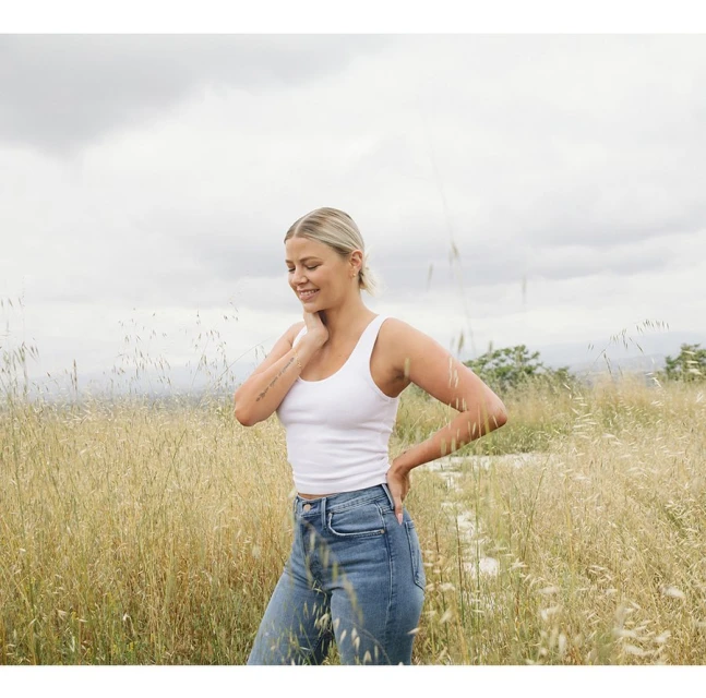 Young woman in a white tank top and blue jeans smiles while standing in a golden field beneath an overcast sky, portrait shot.