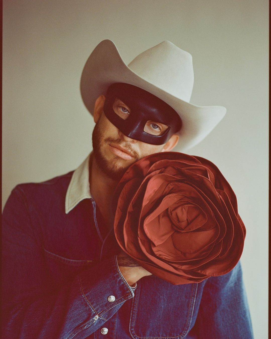 Portrait of a man wearing a black mask and a white cowboy hat, holding a large decorative rose, looking at the camera in a studio setting.