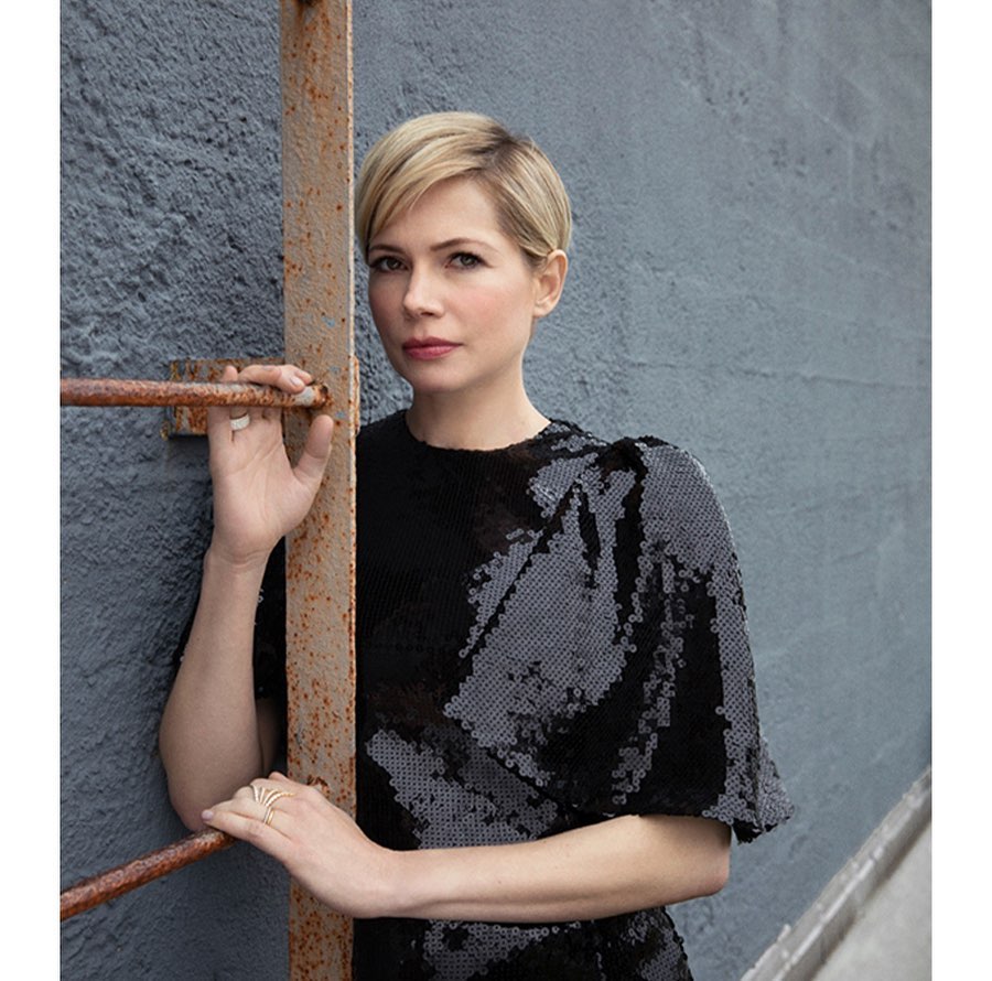 Portrait of fair-skinned adult woman with blonde pixie cut in black sequin dress standing near rusty metal bars and textured wall.