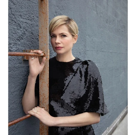 Portrait of fair-skinned adult woman with blonde pixie cut in black sequin dress standing near rusty metal bars and textured wall.