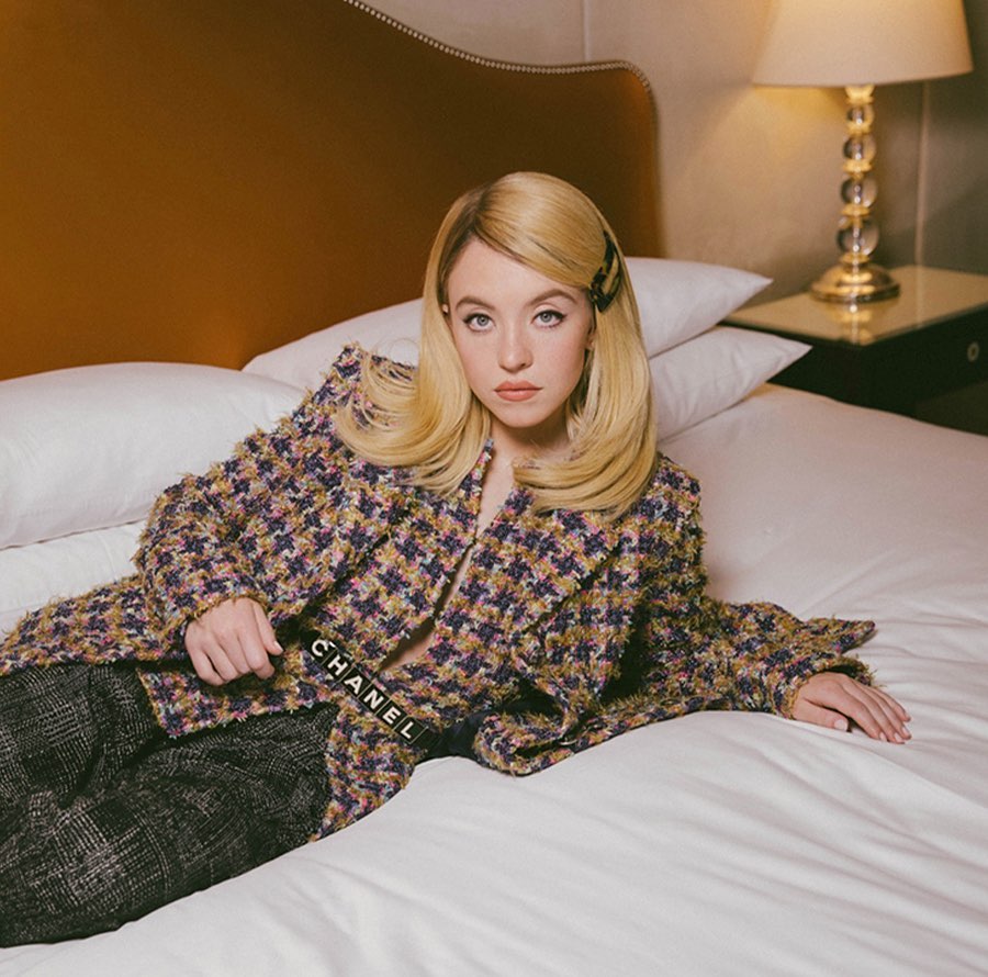 Blonde woman, Sydney Sweeney, lying on a white bed with a Chanel belt and patterned jacket in a hotel room interior.