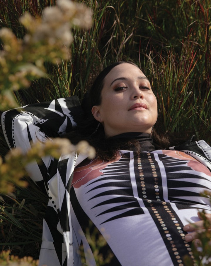A woman with dark hair and patterned top poses while lying in a grassy field with blurred vegetation in an eye-level outdoor shot.