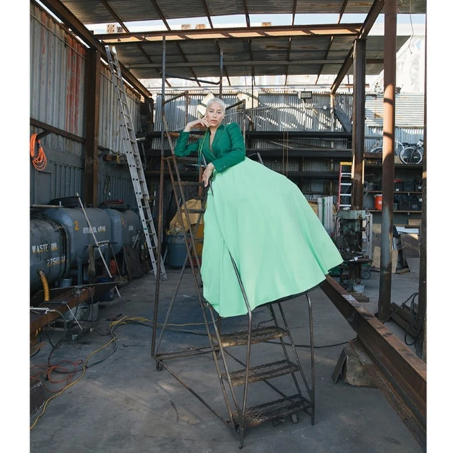 A woman in a green dress and jacket poses on a metal step ladder in an industrial building with tanks and equipment.
