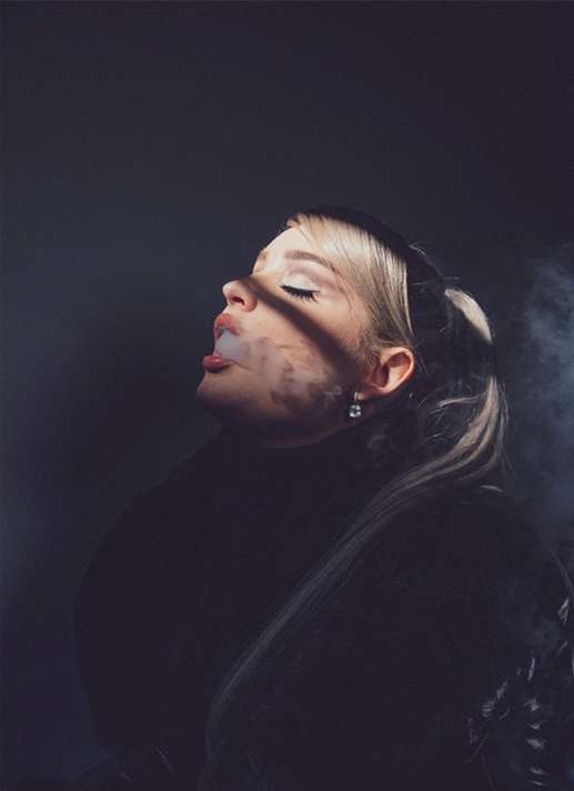 Close-up portrait of a young adult female with blonde hair in a ponytail, smoking and wearing a black turtleneck, against a grey background.