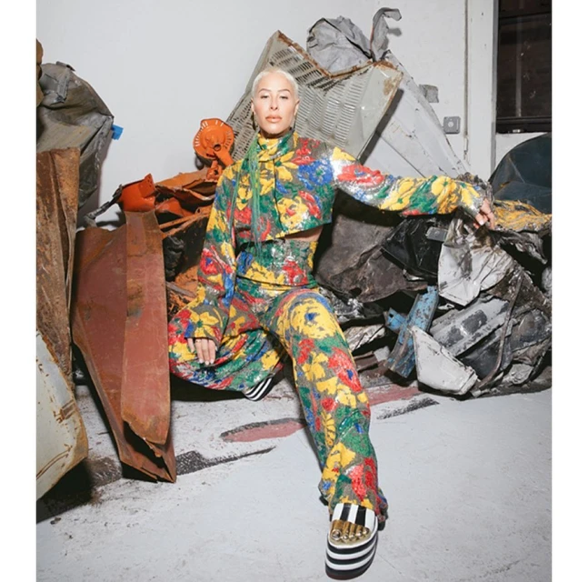 Adult female in a floral crop top and pants poses indoors next to scrap metal, wearing striped platform shoes, with fair skin and short hair.