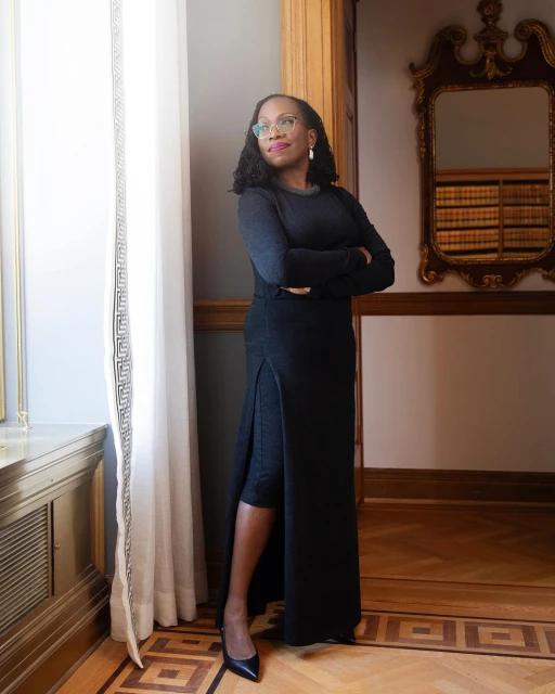 Full-length shot of a Black woman in a long dark dress with a slit, standing indoors near a window with ornate trim and a mirror.