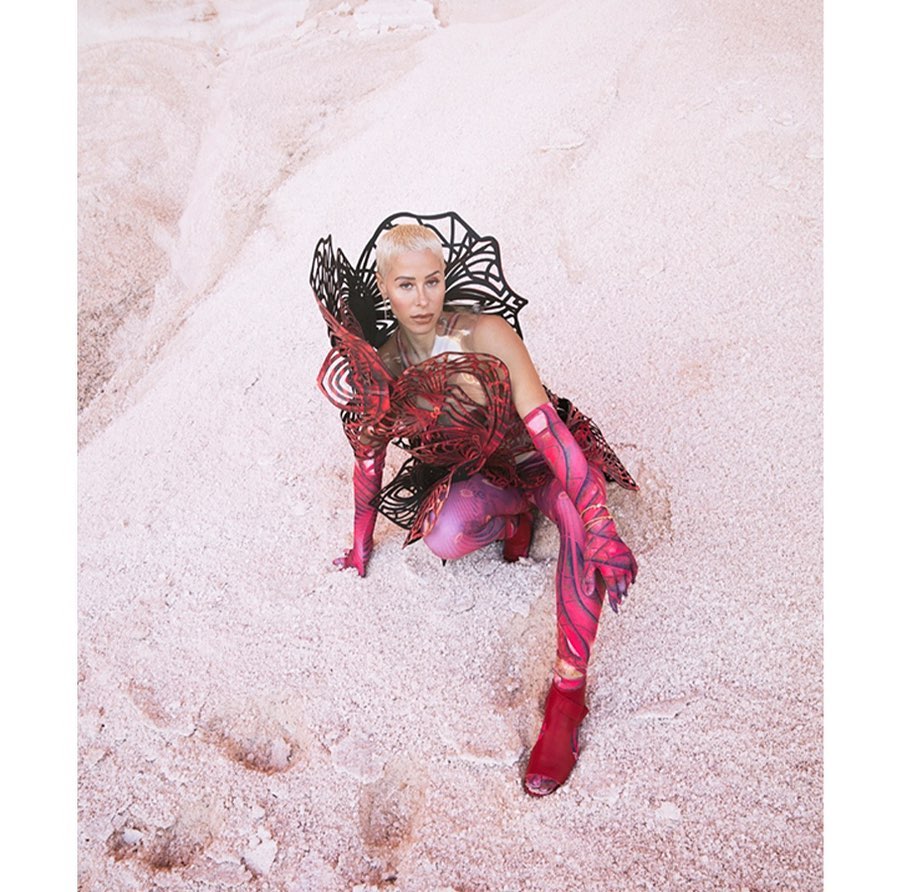 A woman in sculptural fashion crouches on a pink sandy surface, looking directly at the camera in a striking and artistic portrait.