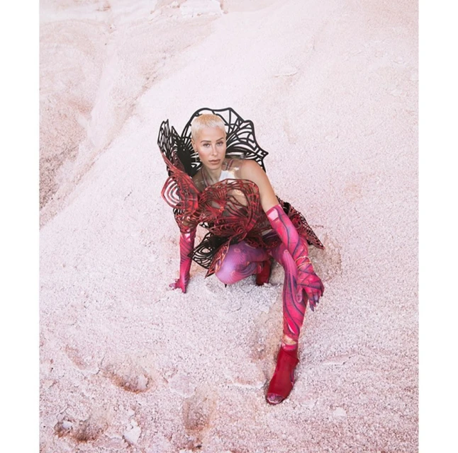 A woman in sculptural fashion crouches on a pink sandy surface, looking directly at the camera in a striking and artistic portrait.