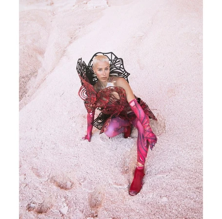 A woman in sculptural fashion crouches on a pink sandy surface, looking directly at the camera in a striking and artistic portrait.