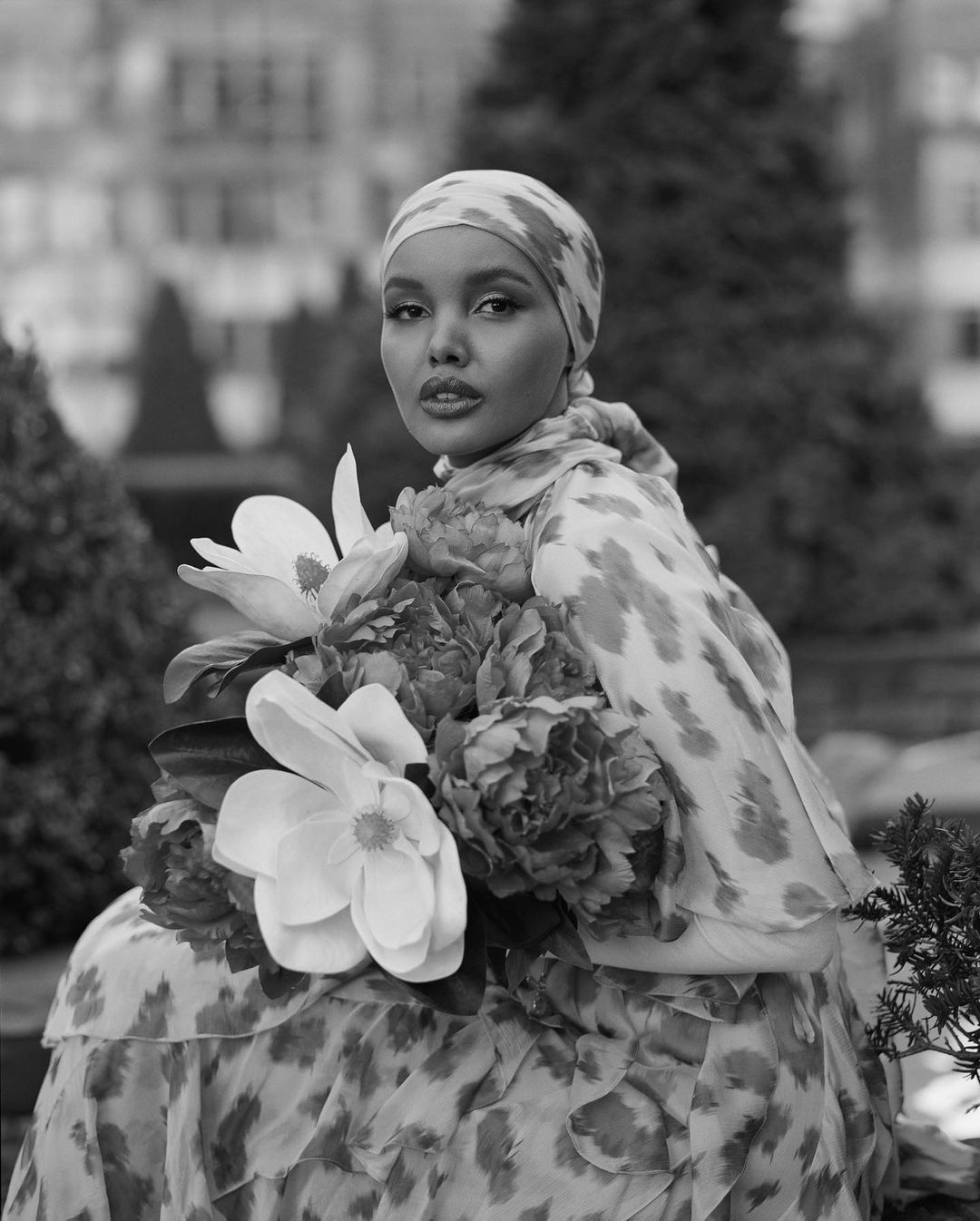 Black and white portrait of a woman in a patterned headscarf and dress, surrounded by large white and dark peonies in an outdoor setting.