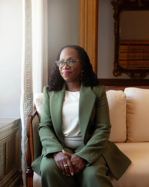 Professional portrait of Ketanji Brown Jackson wearing an olive-green suit, seated on a cream couch near a window in an indoor setting.
