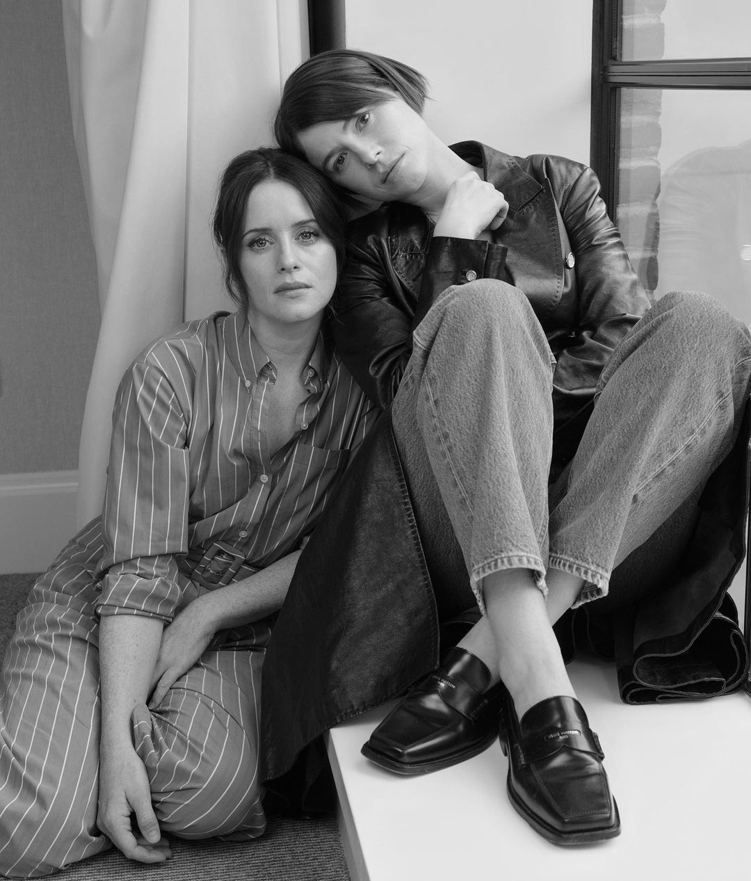 Two women in a black and white portrait. One wears a striped shirt, the other a jacket and jeans, indoors, sitting near a window.