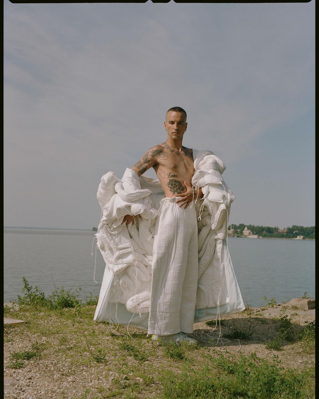 Adult man in white pants stands near water wearing puffy white jacket with tattoos on arms and torso in an outdoor daytime setting.