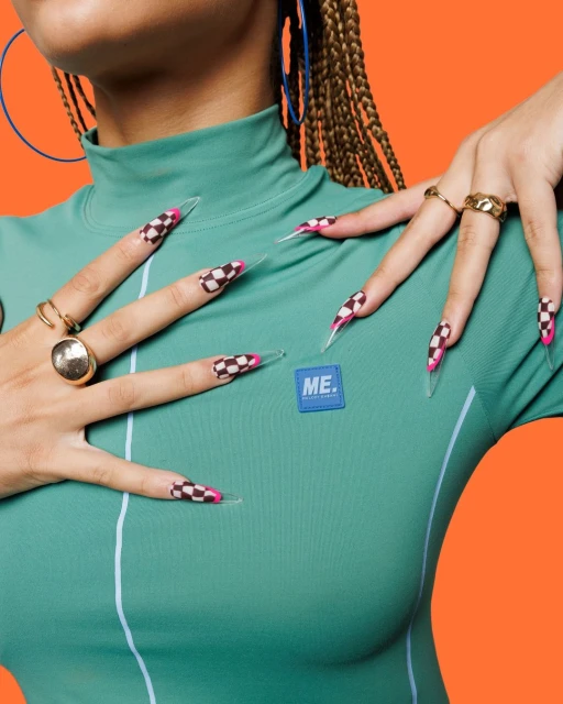 Close-up studio shot of a female torso with long, checkered-pattern nails, wearing a teal shirt against an orange background.