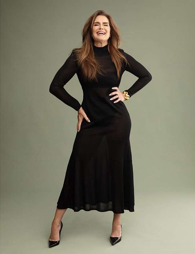 Brooke Shields smiling in a long black dress and high heels against a solid greyish green background photographed by Celeste Sloman
