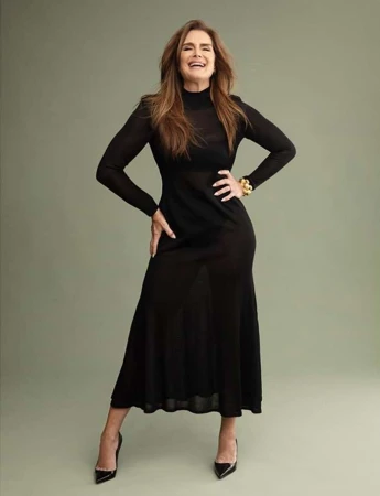 Brooke Shields smiling in a long black dress and high heels against a solid greyish green background photographed by Celeste Sloman