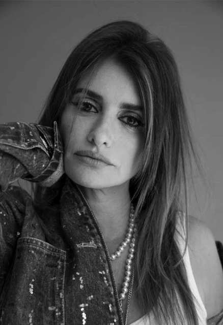 Black and white portrait of a woman in a denim jacket with a pearl necklace, resting her face on her hand, with neutral expression.