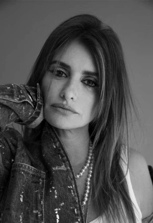 Black and white portrait of a woman in a denim jacket with a pearl necklace, resting her face on her hand, with neutral expression.