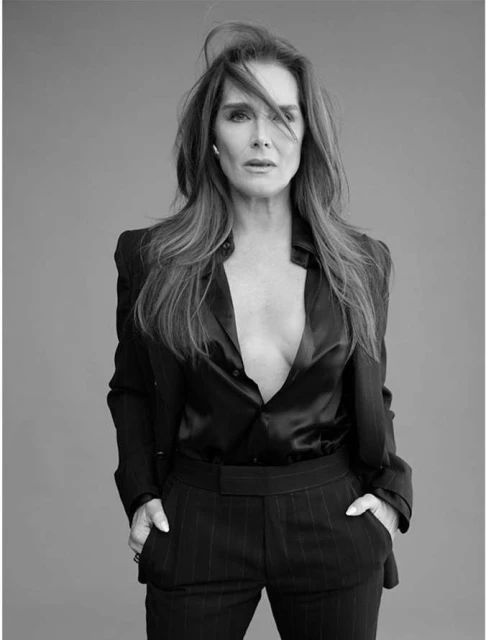 Black and white portrait of a woman in a pinstripe suit, with long brown hair and hands in her pockets against a grey backdrop.