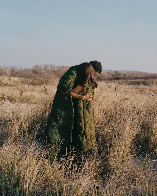 Two people wrapped in a quilt embrace in a field of dry grass under a clear sky, creating a heartwarming outdoor scene with natural tones.