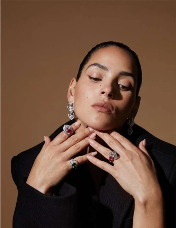 Close-up portrait of a woman wearing statement diamond rings and earrings, posing with hands near her chin against a neutral background.