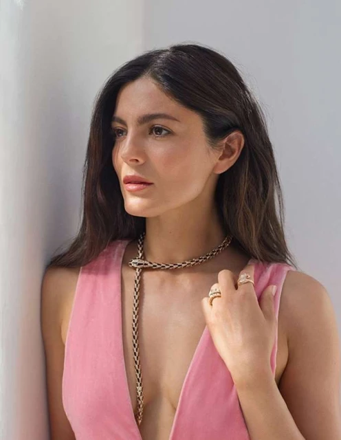 Portrait of a brunette woman wearing a pink dress and gold snake necklace, posing against a white wall in soft, natural light.