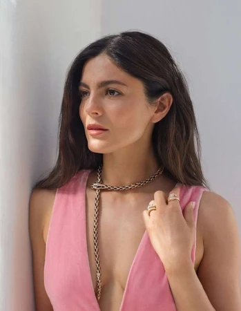 Portrait of a brunette woman wearing a pink dress and gold snake necklace, posing against a white wall in soft, natural light.