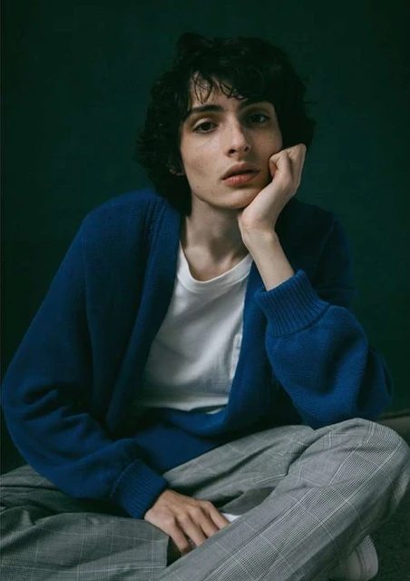 A young adult male with dark curly hair sits indoors wearing a blue cardigan, white t-shirt, and gray checkered pants against a dark background.